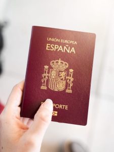 Close-up unrecognizable female hand holds Spanish European passport with unfocused luggage on background. Sunlight and flares. Travel concept.